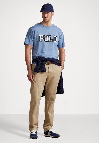 Tricou bleu deschis cu "POLO" scris cu litere îngroșate, pantaloni bej, pulover bleumarin legat în talie, șapcă bleumarin și adidași negri cu accente albe.