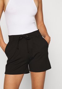 Shorts noirs en tissu tissé avec une taille élastique, un cordon de serrage et des poches latérales. Le tissu semble lisse avec une légère élasticité.