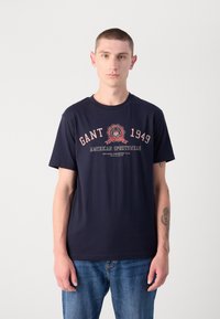 Tricou din bumbac bleumarin cu guler rotund și mâneci scurte, având un design grafic central care afișează "GANT 1949 American Sportswear."
