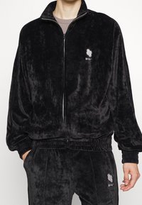 Zwarte velours jas met ritssluiting, hoge kraag, elastische manchetten en tailleband; logo op de borst. Zachte textuur en ontspannen pasvorm.