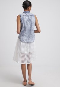 Femme aux cheveux foncés portant une chemise sans manches en denim délavé à l'acide et une jupe blanche à rayures arrivant aux genoux, chaussée de ballerines couleur nude, debout devant un fond uni clair.