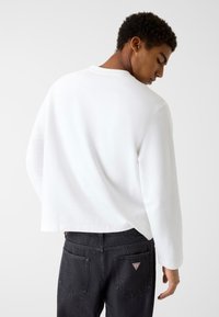 Biała koszula z długim rękawem i okrągłym dekoltem, wykonana z prążkowanej tkaniny, noszona z czarnymi dżinsami z małym trójkątnym logo na tylnej kieszeni.