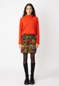 Rode gebreide pullover met een hoge kraag en knopdetails, gecombineerd met een bloemenpatroon minirok met kleurrijke bloemen en zwarte panty’s.