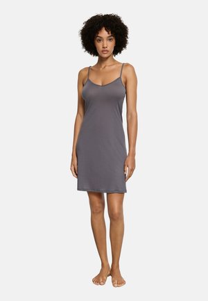 Kobieta o kręconych włosach, stojąca boso, ubrana w szarą, bezrękawną, sięgającą kolan sukienkę na ramiączkach, na tle jednolitego białego tła.