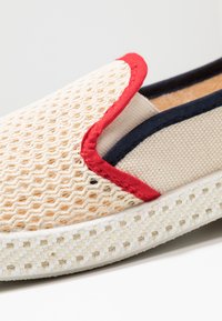 Slip-on schoen met een beige mesh bovenwerk, rode en marineblauwe accenten, een gestructureerde zool en een geweven ontwerp. Zachte voering van stof en stevige constructie.