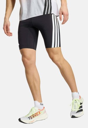 Uomo che indossa pantaloncini sportivi neri con strisce bianche e scarpe da corsa Terrex bianche con lacci giallo neon, in piedi con gambe divaricate.