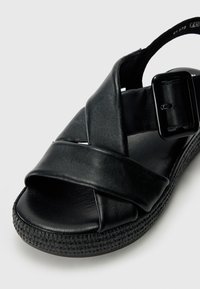 Sandale en cuir noir avec une conception de lanières croisées, boucle ajustable et semelle texturée avec des rainures subtiles pour une meilleure adhérence.
