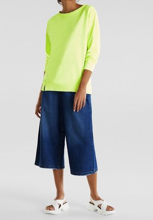 Neon-gelbes Langarmshirt mit lockerer Passform, kombiniert mit weiten, mittelblauen Denim-Culottes und weißen Slipper-Sandalen.