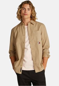 Camicia color khaki con bottoni, dotata di una tasca sul petto e un patch con logo. Indossata sopra una maglietta rosa chiaro, con le maniche arrotolate.