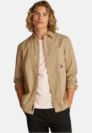 Camicia color khaki con bottoni, dotata di una tasca sul petto e un patch con logo. Indossata sopra una maglietta rosa chiaro, con le maniche arrotolate.