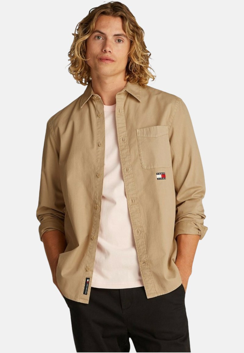 Camicia color khaki con bottoni, dotata di una tasca sul petto e un patch con logo. Indossata sopra una maglietta rosa chiaro, con le maniche arrotolate.