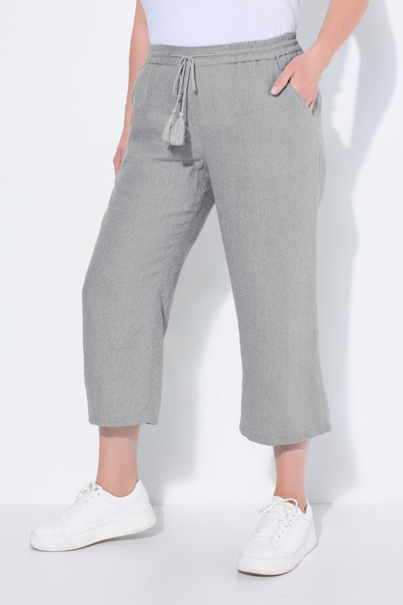 Person trägt graue, gekürzte Hosen mit Kordelzug in der Taille und weiße Sneakers, Hand in der Tasche ruhend, vor weißem Hintergrund.