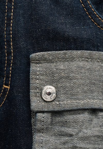 Κοντινή λήψη μπλε τζιν παντελονιού με γκρι ύφασμα πτυχής τσέπης, στερεωμένη με ασημένιο κουμπί που έχει χαραγμένο το όνομα της μάρκας.