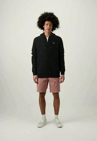 Black zip-up hoodie with drawstrings, features a small logo on the left chest. Paired with light pink shorts and white sneakers.