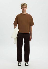 Bruine korte mouwen T-shirt, zwarte rechte broek, witte trui over de arm gedrapeerd, zwarte schoenen met witte accenten, neutrale achtergrond.