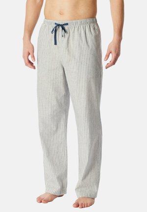 Man die lichtgrijze pyjamabroek draagt met witte verticale strepen en een donkerblauwe trekkoord, staand op blote voeten tegen een witte achtergrond.