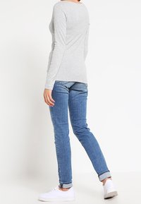 Person som står på sidan iförd en ljusgrå långärmad tröja, blå uppvikta jeans och vita sneakers mot en enkel vit bakgrund.