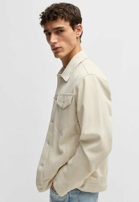 Beige denimjacka med spetsig krage, två bröstfickor och knappstängning fram. Mjuk textur, avslappnad passform och synliga sömnadsdetaljer.