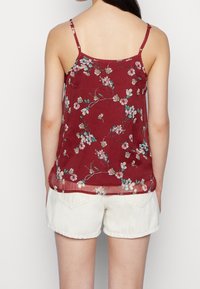 Top sin mangas de color burdeos con tirantes finos; presenta motivos delicados de flores en blanco y azul. Combinado con pantalones cortos de un color claro.