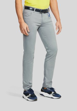 Mann trägt hellgraue Slim-Fit-Hose, schwarzen Gürtel mit blauem Karabiner, gelbes Hemd und dunkelblaue und weiße Sneakers, steht.