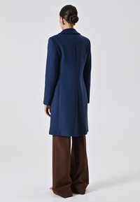 Donna con i capelli raccolti a bun che indossa orecchini d'oro, un cappotto blu navy lungo fino al ginocchio e pantaloni larghi marroni, in piedi su uno sfondo chiaro.