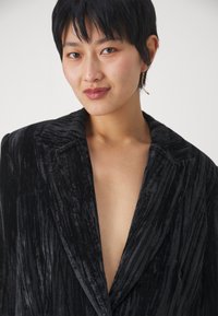 Sort fløjl blazer med en tekstureret, ribbet overflade og dyb V-udskæring. Har brede skuldre og en skræddersyet pasform, der viser et minimalistisk design.