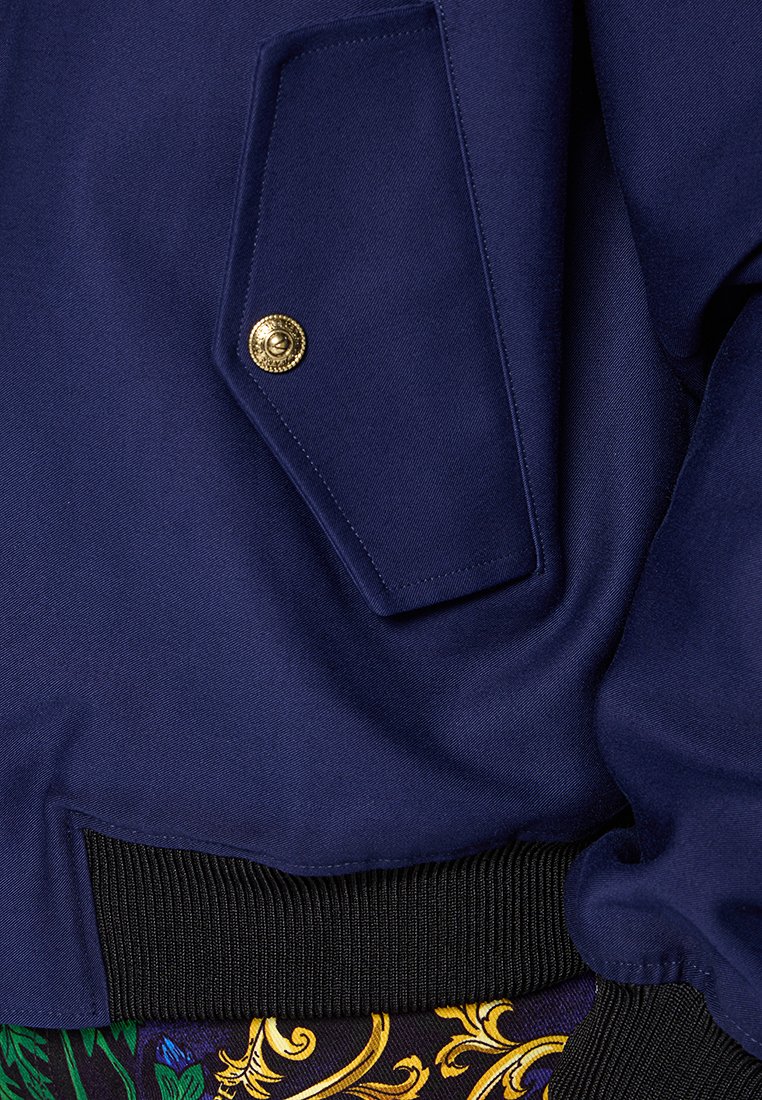 Marineblaue Jacke aus strukturiertem Stoff, mit einer auffälligen Klappentasche und goldenen Knopfdetails; geripptem schwarzem Saum.