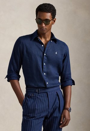 Mann iført mørkeblå skjorte med knapper og oppsatte ermer, stripete marineblå bukser, solbriller og armbåndsur, står foran en beige bakgrunn.