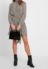 Vestido estampado con mangas largas, cintura ceñida y cuello alto. Bolso de mano negro con efecto cocodrilo y asa verde. Botines negros.