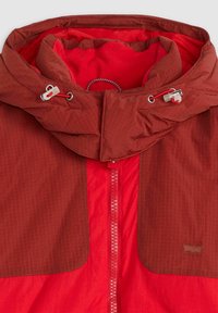 Giacca rossa con cappuccio, pannelli anteriori marroni, chiusura frontale con zip, cordini regolabili e morbida fodera in pile all'interno del cappuccio.