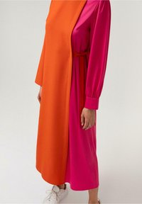 Femme portant une longue robe rose avec des manches froncées et un manteau asymétrique orange sans manches, associée à des baskets blanches.