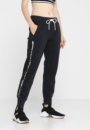 Kvinde iført sorte Under Armour træningsbukser med hvidt logotekst på benet, kombineret med sorte og hvide sportssko, stående med hænderne i lommerne.