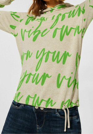 Pull beige avec texte cursif vert vif et détail à cordon, porté avec un jean bleu foncé sur une personne aux cheveux bruns mi-longs.
