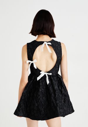 Robe noire avec un dos nu, ornée de trois accents en ruban blanc. Confectionnée en tissu texturé avec un corsage ajusté et une jupe évasée.