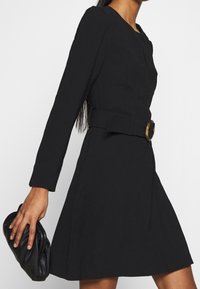 Černé šaty s páskem, dlouhými rukávy, ušitým korzetem, rozšířenou sukní a velkým kruhovým knoflíkem v pase. Hladká textura, minimalistický design.