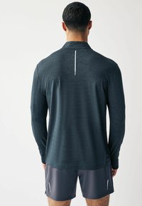 Langärmeliges Sportshirt aus dunkelgrauem Stoff mit strukturiertem Finish. Enthält einen subtilen vertikalen reflektierenden Akzent auf der Rückseite. Getragen mit Shorts.