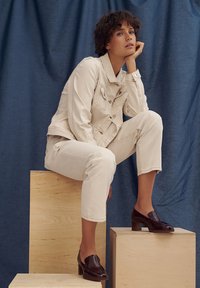 Beige denim jacket with snap buttons, paired with matching beige cropped pants. Dark brown heeled loafers complete the outfit. Blue backdrop.