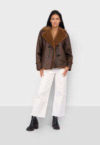 Brązowa kurtka z owczej wełny z dużym kołnierzem, zapinana na podwójne guziki, w połączeniu z białymi spodniami o szerokich nogawkach i czarnymi botkami za kostkę.