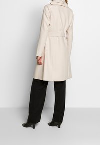 Gabardina beige con cintura ceñida, cuello alto y mangas largas. La tela parece suave; la gabardina llega hasta la mitad del muslo, estilizada sobre pantalones negros.