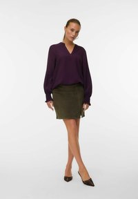 Haut violet à manches longues avec un décolleté en V et des poignets froncés, associé à une mini jupe en daim verte avec une fente sur le côté. Ballet flats pointus marron.