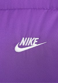 Bílé vyšívané logo Nike na strukturované fialové tkanině s hexagonálním vzorem.