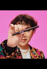 Un stylo eyeliner noir tenu par une main avec des ongles multicolores, portant une veste à motifs colorés. L'arrière-plan est d'un rose uni.