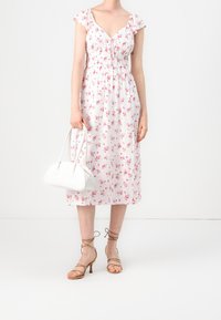 Nainen, joka on pukeutunut valkoiseen midi-mekkoon, jossa on vaaleanpunaisia kukkakuvioita, vaaleanruskeisiin nauhakenkiin ja hän pitää valkoista käsilaukkua yksinkertaista valkoista taustaa vasten.