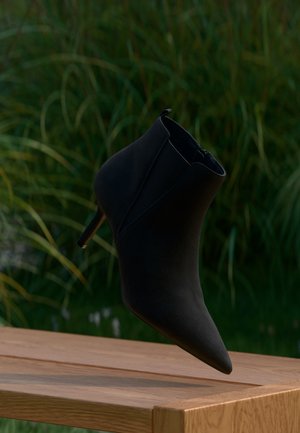 Svart ankelstøvel i semsket skinn med spiss tå og høye hæler, plassert over en treflate med grønt løvverk i bakgrunnen.