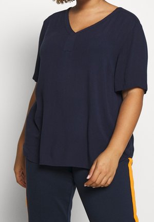 Femme portant un haut ample bleu marine à col en V et manches courtes, et un pantalon bleu marine avec une bande orange sur le côté.