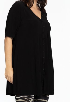 Woman wearing a loose black button-up tunic with short sleeves and zebra-patterned leggings against a white background.