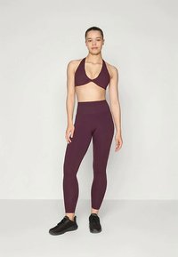 Burgundy sports bra with twisted front design paired with matching high-waisted leggings. Model wearing black athletic shoes, standing upright.