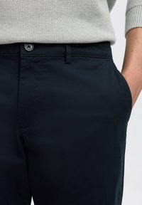 Nahaufnahme einer Person, die dunkelblaue Hose mit Knopf und eine Hand in der Tasche trägt, kombiniert mit einem hell strukturierten Pullover.