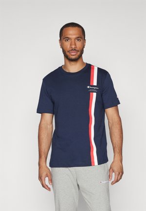 Camiseta de algodón azul marino con una atrevida raya vertical roja y blanca; logo en el pecho; cuello redondo acanalado; combinada con pantalones de chándal grises.