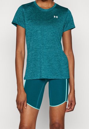 Persona vistiendo una camiseta deportiva de manga corta color azul verdoso y pantalones cortos de bicicleta del mismo color con ribete azul claro, de pie contra un fondo liso.